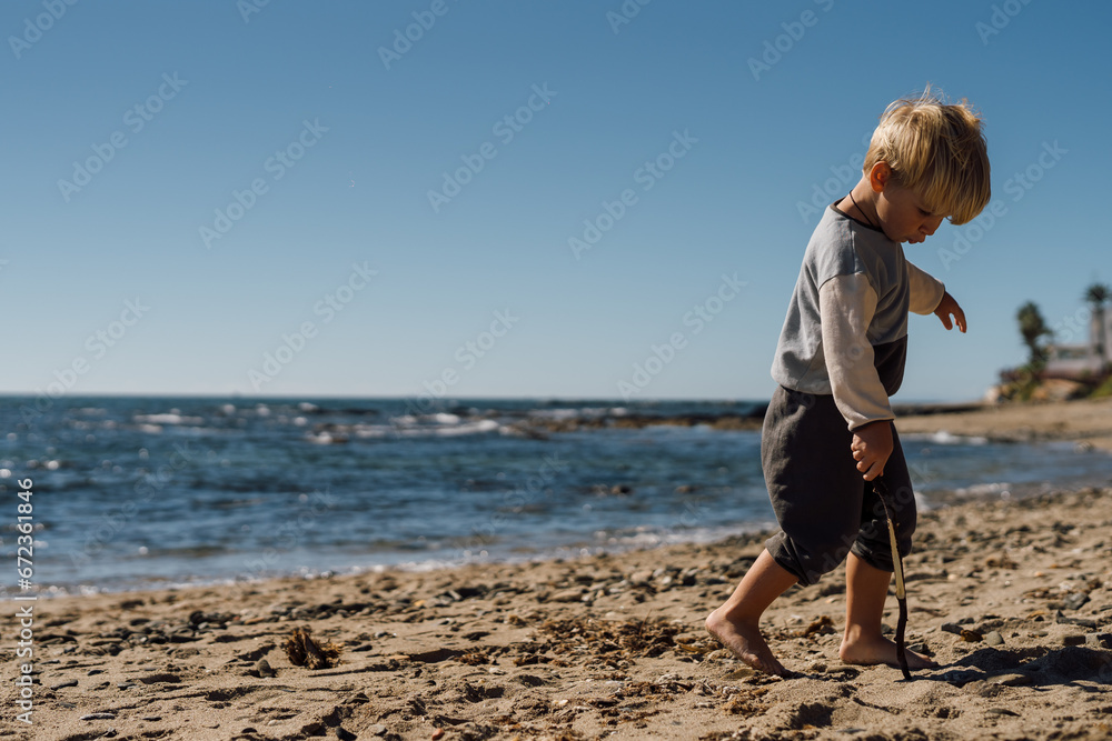 A beautiful three-year-old blond boy walks barefoot on the sandy shore by the sea.
