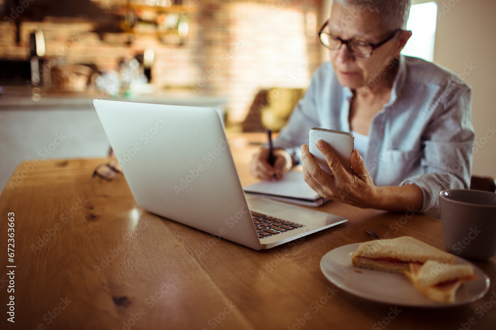 Elderly woman holding smartphone writing notes with laptop at home Elderly woman holding smartphone writing notes with laptop at home
