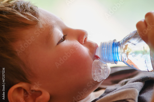 A child drinks water from a blue plastic bottle on the background of a window. Toddler baby with a bottle of clean water in his hands. Kid aged two years