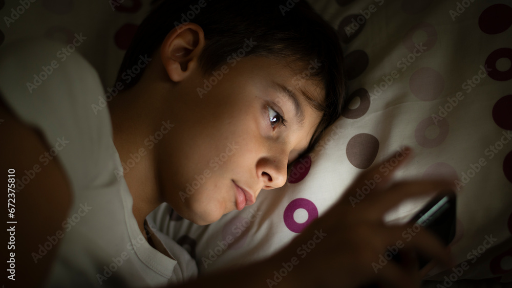 Child inside social networks with his smartphone from bed at night before sleeping. Concept of kids using social networks and their dangers without parental control at night from their room - obrazy, fototapety, plakaty 