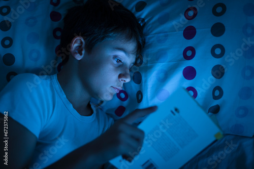 boy uses smathphone hidden in the book at night in bed before falling asleep. Concept of the problem of mobile phone use in children. Boy with phone inside a book illuminated by screen at night photo