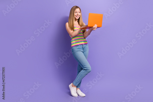 Full length photo of lovely teen blonde lady orange netbook typing wear trendy striped garment isolated on purple color background