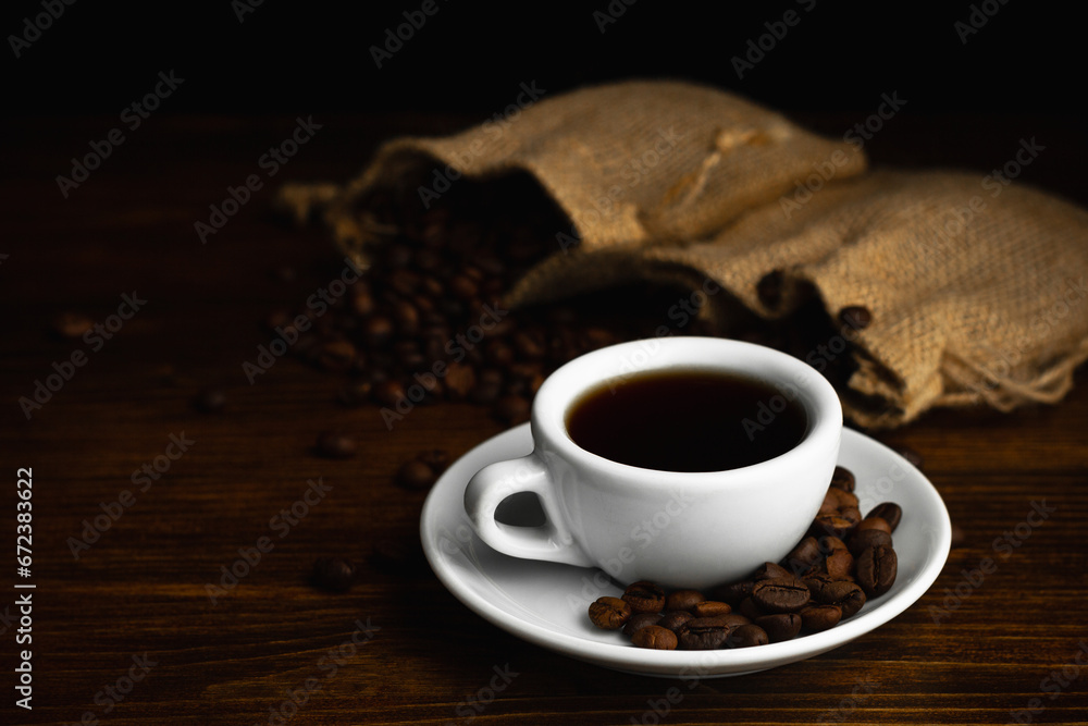 Hot coffee in a white coffee cup and many coffee beans placed around on a wooden table in a warm, light atmosphere, on dark background, with copy space. Hot coffee in a white coffee cup and many coffee beans placed around on a wooden table in a warm, light atmosphere, on dark background, with copy space.