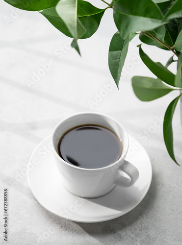 A white cup of black aromatic coffee on a light background with green branch and shadow. Morning energy drink concept.