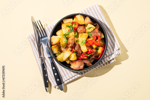 Fried pork meat with potatoes, onions and peppers. National homemade Georgian dish ojakhuri in a black bowl on a yellow background with fork, knife and shadows. photo