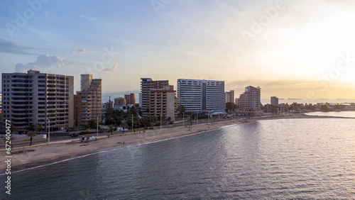 Drone photos of the City of Lechería on a sunset, in which you can see buildings, sea, people bathing on the beach, and passers-by. Playa los Canales Boulevard