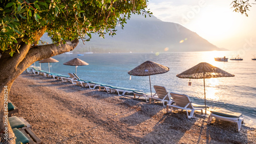Fototapeta Naklejka Na Ścianę i Meble -  Empty wooden sun loungers and umbrellas on the beach, behind them the mountain covered with green forests, no people, Clean and clear Aegean sea Muğla - Turkey