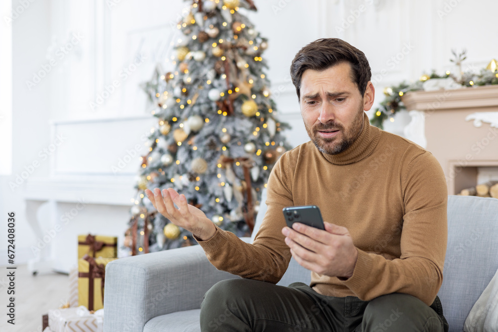 Upset unhappy man sitting near Christmas tree on winter day, got bad news on phone, celebrating new year and Christmas, unhappy reading, using app on smartphone, living room at home on sofa