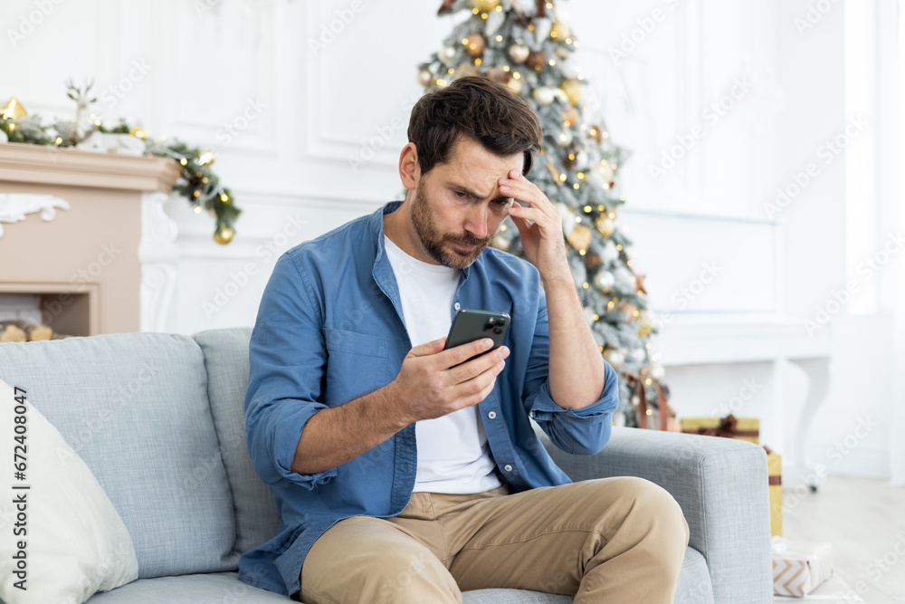 Upset unhappy man sitting near Christmas tree on winter day, got bad news on phone, celebrating new year and Christmas, unhappy reading, using app on smartphone, living room at home on sofa Upset unhappy man sitting near Christmas tree on winter day, got bad news on phone, celebrating new year and Christmas, unhappy reading, using app on smartphone, living room at home on sofa