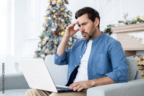 Frustrated sad and unhappy man sitting on sofa in living room near Christmas tree, businessman working remotely on New Year holidays, looking at laptop confused