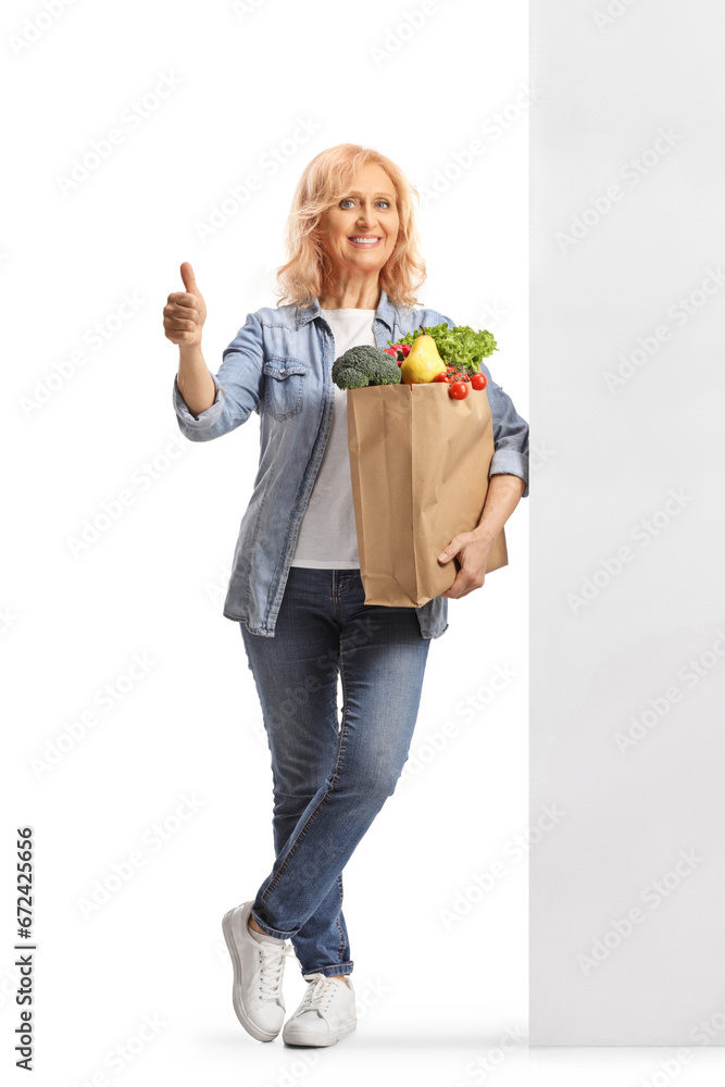 Full length portrait of a mature woman with a grocery bag standing against a wall and gesturing thumbs up Full length portrait of a mature woman with a grocery bag standing against a wall and gesturing thumbs up