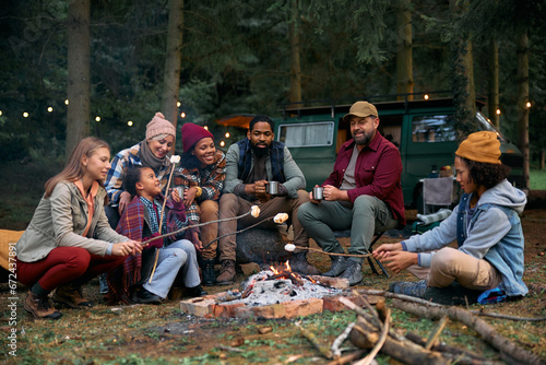 Multiracial families roasting marshmallows while gathering around campfire in woods. photo