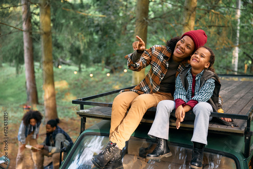 Happy black mother and daughter observing nature while sitting on family camper trailer's roof.