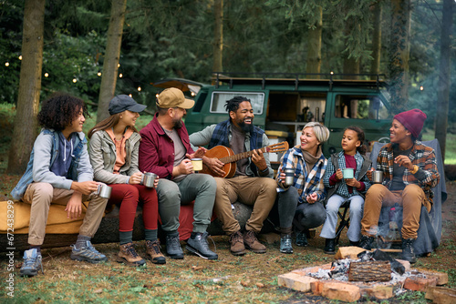 Happy black man plays acoustic guitar while camping with his family and friends in nature.