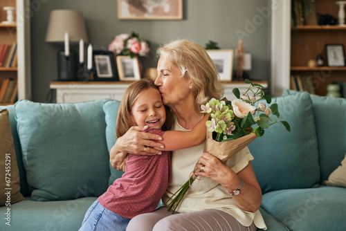 Happy grandma hugging and kissing granddaughter thanking for present, holding flower bouquet