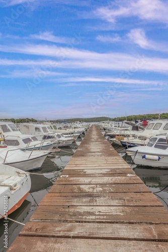 View along a wooden jetty in a harbor with docked ships in front of blue skies
