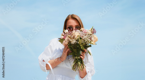 Joyful Young Woman Holding a Bouquet of Flowers on a Beach with Blue Sky Background