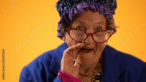 Closeup funny portrait of smiling happy crazy toothless grandmother with wrinkled skin puts hand to mouth to tell a secret isolated on yellow background studio photo