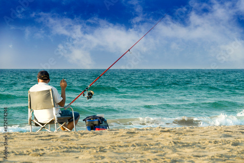 An elderly man sits on the seashore, smokes and fishes with a fishing rod.