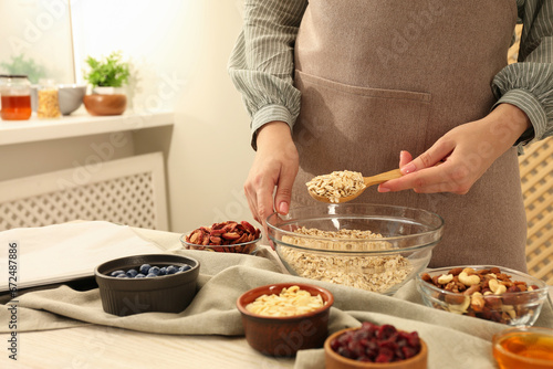 Making granola. Woman putting oat flakes into bowl at table in kitchen  closeup