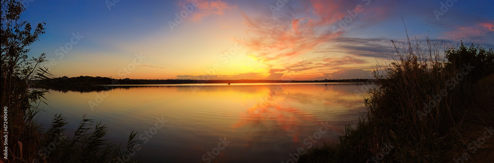 Stunning sunset on the shore of a lake with reeds in the foreground. panorama