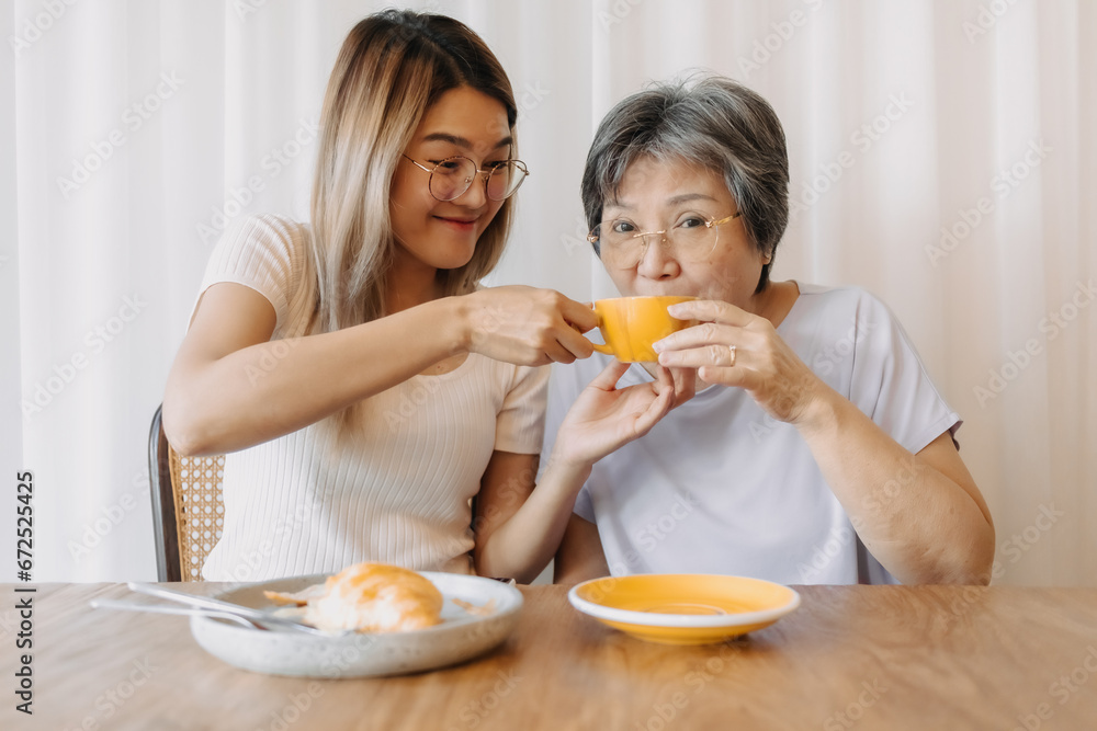 Happy Asian old woman mother and adult daughter wear glasses, eating croissant bakery and drinking coffee tea at cafe, holding cup and spending happy life time together over white curtain.