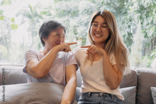 Happy Asian Thai mother and daughter sitting on sofa, smiling and laughing with love, playing and pointing fingers to each other, spending good time, family life relationship.