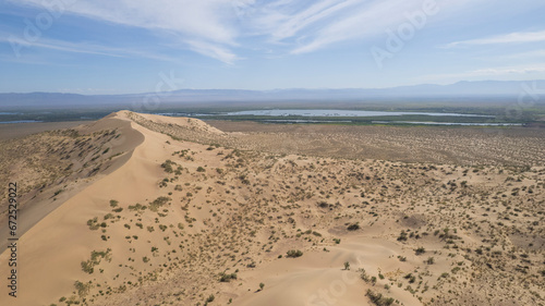 A large sand dune in the middle of the steppe. Drone view of a huge pile of sand. Tourists are walking, enjoying the view. A river runs in the distance and grass grows. The sky with white clouds
