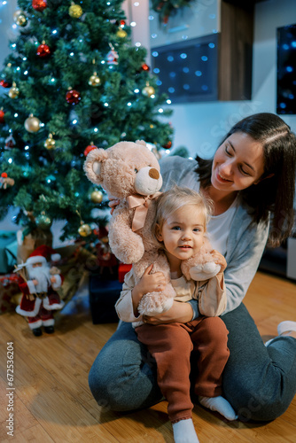 Mom with a little girl and a teddy bear sits on the floor near the Christmas tree
