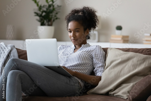 Happy Black student girl studying from home, resting on comfortable couch, using laptop, watching webinar, virtual training, online learning seminar, making video call to teacher