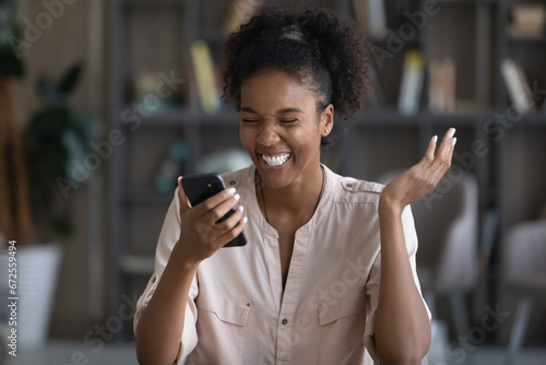 Happy excited young Black woman reading text on mobile phone screen, getting amazing surprising good news, celebrating success, achieve, result. Overjoyed gen Z student girl using smartphone, laughing