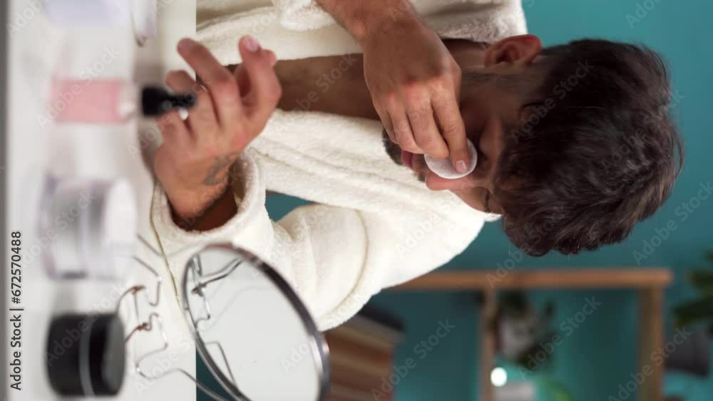 Handsome Arabic young man looking into mirror uses cotton pad for cleans his face, removing makeup before shower, skin care at home concept
