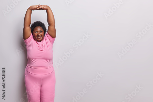 Sporty lifestyle. Indoor shot of young happy smiling plump darkskinned woman standing on left isolated on white background in pink tracksuit doing stretching exercise with blank space for promotion photo