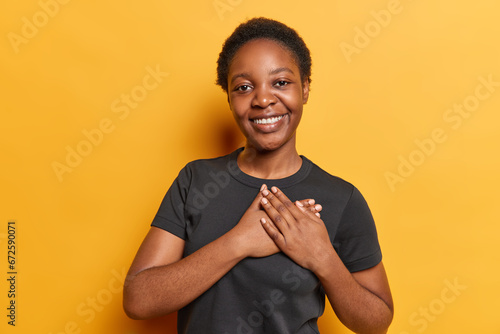 Sign of affection and appreciation. Dark skinned millennial girl with short curly hair presses hands to heart expresses gratitude to you for heartwarming words wears casual black t shirt isolated