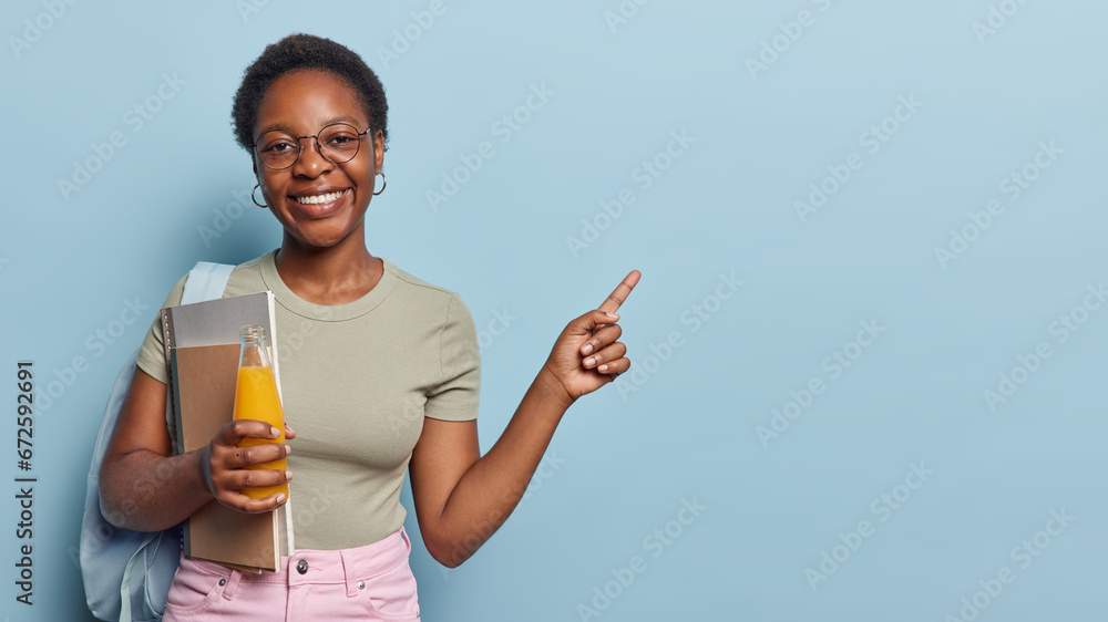Horizontal shot of cheerful dark skinned female college student poses with spiral notepads and bottle of fresh orange juice indicates at blank space over blue studio background smiles broadly. Horizontal shot of cheerful dark skinned female college student poses with spiral notepads and bottle of fresh orange juice indicates at blank space over blue studio background smiles broadly.