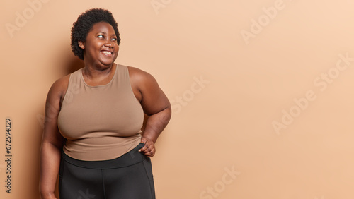 Fotografie Horizontal shot of positive curly haired African woman has excess weight dressed in t shirt and leggings concentrated aside smiles positively goes in for sport isolated over brow background