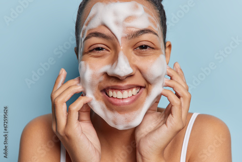 Close up shot of cheerful woman washing face with foam cleanser smiles gladfully shows white teeth cleans skin stands bare shouldered isolated over blue background. Pampering procedures at home photo