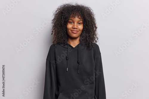 Positive curly haired young African woman dressed in black sweatshirt has satisfied expression smiles gently wears black casual sweatshirt isolated over white background. People emotions concept © wayhome.studio 