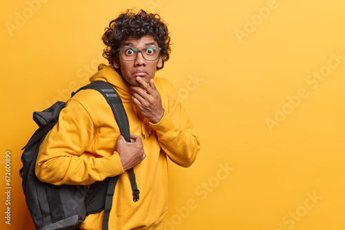 Horizontal shot of shocked Hindu man keeps hand on chin stares impressed at camera hears something astonishing dressed in casual sweatshirt carries rucksack on shoulder isolated on yellow wall