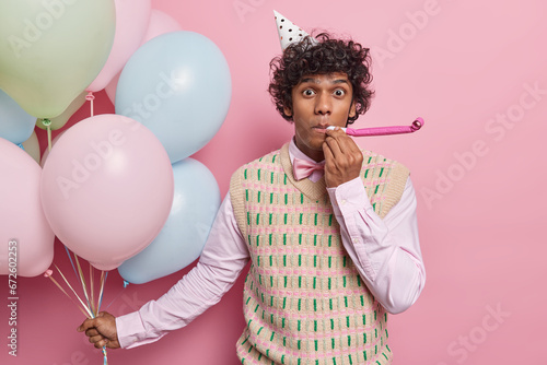 Handsome curly haired Hindu man blows into party horn wears cone hat formal shirt and vest holds bunch of inflated balloons isolated over pink background. People holiday and celebration concept photo