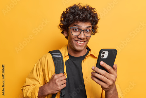 Studio shot of cheerful Hindu man with dark curly hair uses smartphone smiles gladfully poses with rucksack dressed in casual clothing isolated over yellow background. People and lifestyle concept