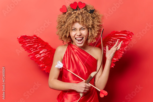 St. Valentines Day celebration. Indoor photo of young pretty excited glad African american woman in cupid costume standing isolated on red background holding bow and arrow with positive emotions photo
