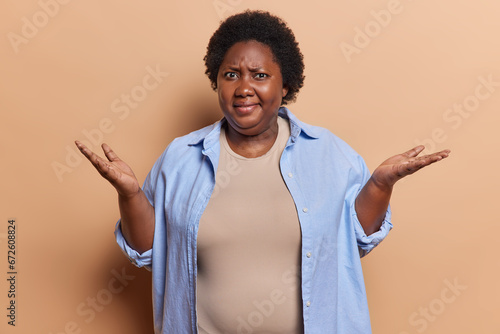 Photo of plump dark skinned woman with curly hair has puzzled displeased expression doesnt know what to do in this situation wears blue shirt isolated over brown background. Hmm I dont know who cares