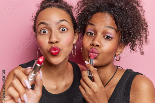 Horizontal shot of two friendly young women use best cosmetic product apply lipstick do makeup to have fabulous look wear black t shirts isolated over pink background. Beauty procedures concept photo