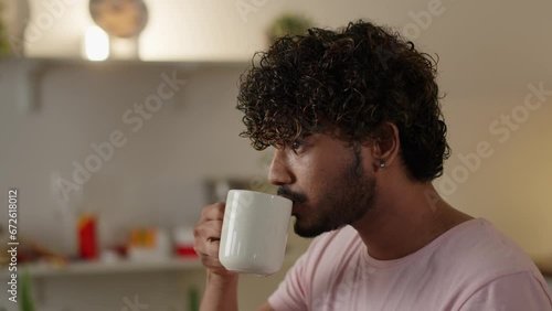 Concept of breakfast. Young guy eating tasty toast and drinking a cup coffee. Man consumes unhealthy bread sandwich with tea at home kitchen. Relaxed student has lunch