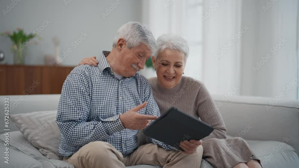 Senior couple man and woman watching something funny in tablet and laughing sitting on couch in living room at home. Family, happy spouses elderly wife and husband hugging, having fun together. Senior couple man and woman watching something funny in tablet and laughing sitting on couch in living room at home. Family, happy spouses elderly wife and husband hugging, having fun together.