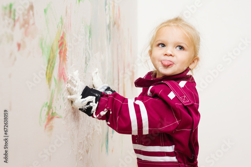 Little blonde girl in overalls playing with white color on wall photo