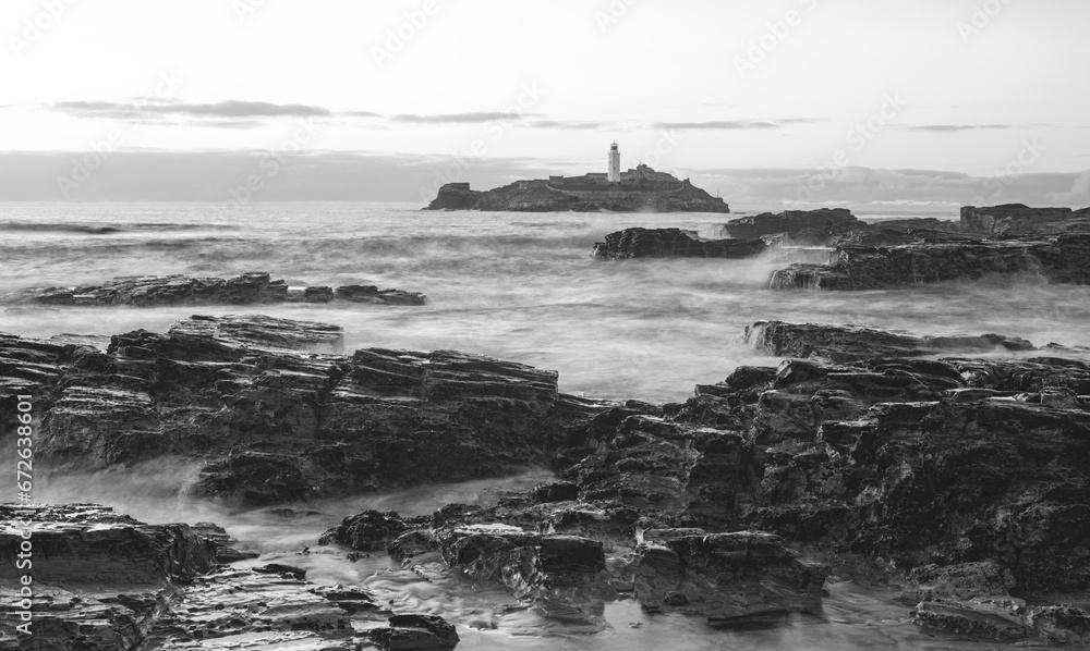 Black and white image of a rocky shoreline with a lighthouse in the background.