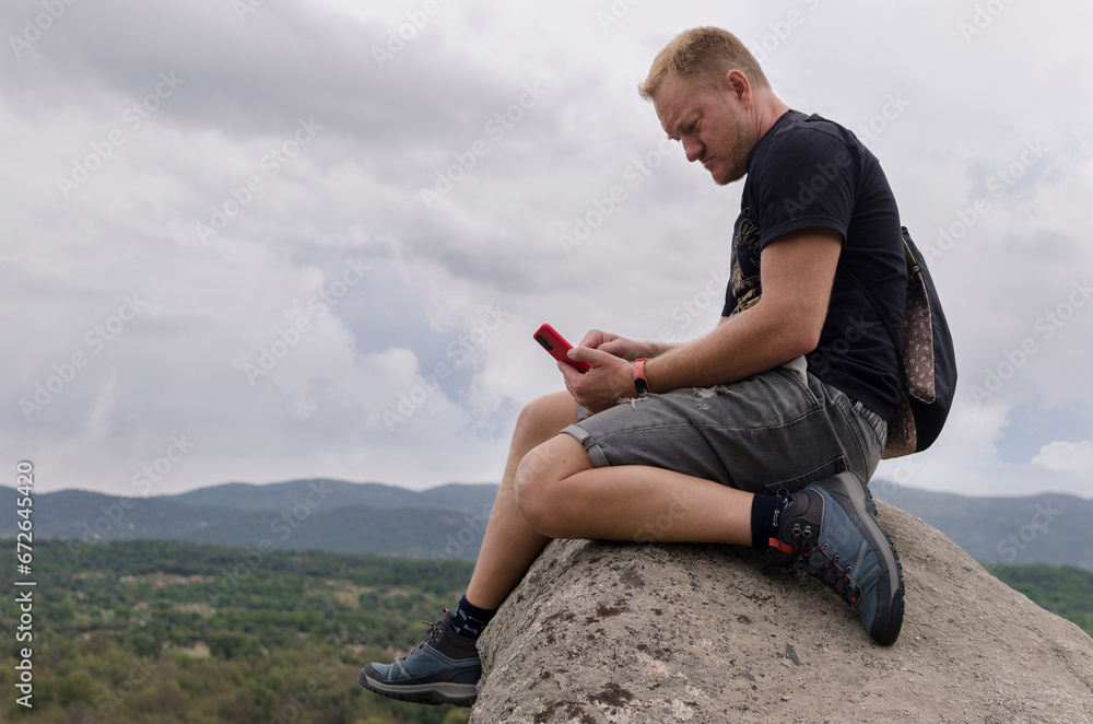 Thoughtful man sitting on a rock on top of mountain peak looking at cell phone screen in hand. Concept of person connected to internet everywhere. Beautiful sky on a cloudy day. Freedom and adventure