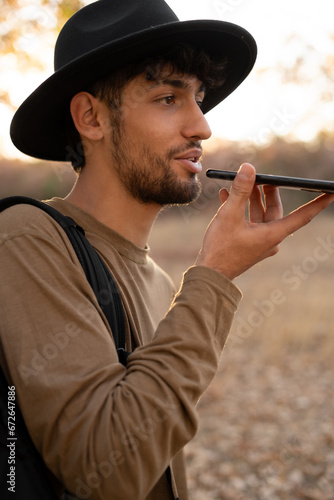 Male traveler wearing hat using a smartphone voice recognition online, walking and talking outdoor. Voice chat bot, conversational AI. © mtrlin